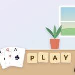 A sunny table with a cup of coffee, a fan of playing cards, wooden letter tiles spelling PLAY and a phone showing a colorful tile puzzle, with a window and a plant behind.